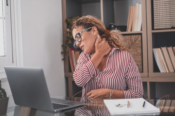 Woman sitting at a table with a laptop in front of her. She is holding her neck and has her eyes closed, as if in pain.