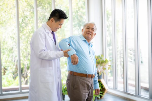 A male doctor stood with a male patient, the doctor is supporting the patient's right arm so it is bent to a right angle.