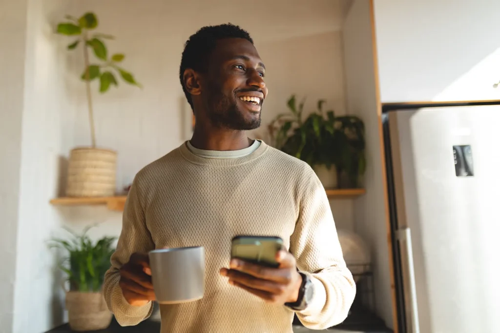 Young man in his kitchen, holding a mug of coffee and his phone.