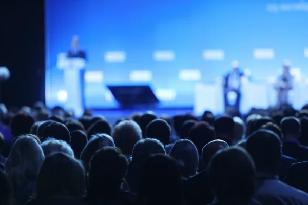 A dark auditorium, looking at a stage where a blurred out individual is talking to a crowd of people
